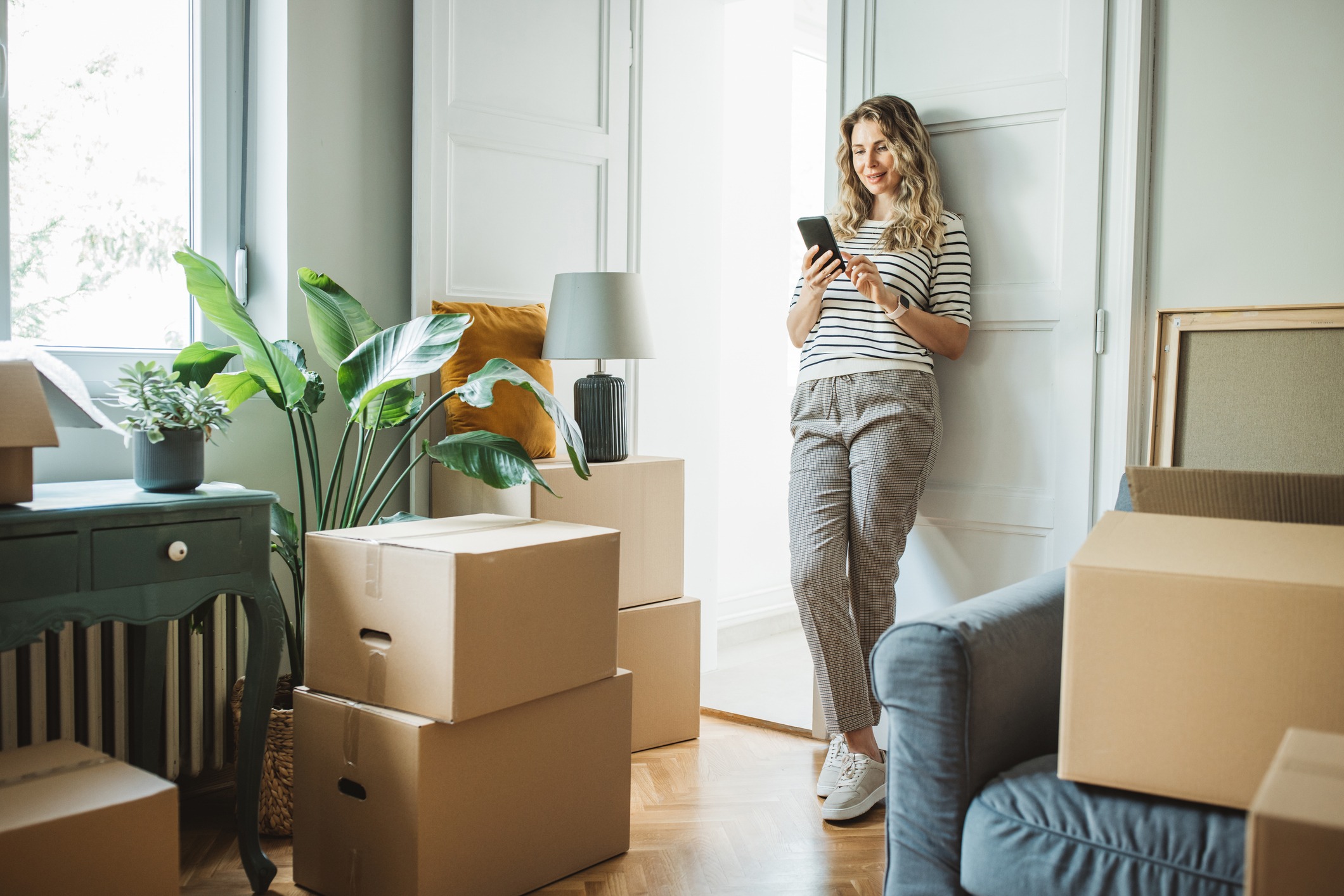 A woman surrounded by moving boxes in her home orders a moving service on her phone.